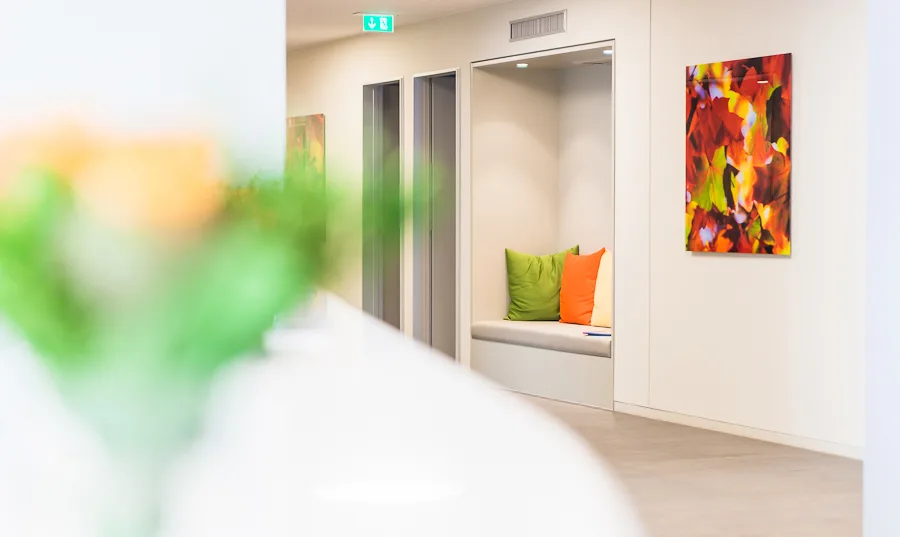 The view shows, leading past a bouquet of flowers standing on a stand, the wall of a corridor of the gynaecological specialist practice for fertility treatment on Berlin's Kurfürstendamm, decorated in cheerful colours. An upholstered bench with back cushions invites you to linger in an alcove.