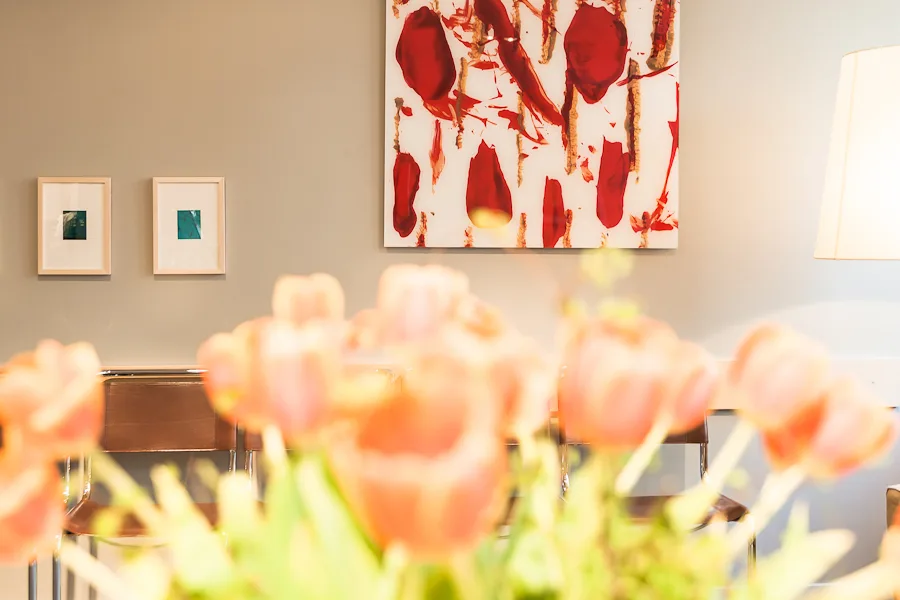 Blick durch ein Blumenbouquet auf eine sandfarbene Wand der Arztpraxis am Kudamm für Kinderwunschbehandlung. Ein großes abstraktes Gemälde und zwei kleinere Bilder werden von einer modernen Stehleuchte erhellt. Mehrere erdfarbene Stühle bieten Platz zum bequemen Sitzen.